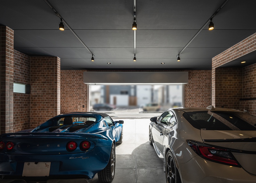 愛車2台をゆったりと格納できるインナーガレージは、まさに大人の秘密基地。<br />
レンガ調の壁面と黒い天井を組み合わせ、重厚感のある落ち着いた空間に仕上げました。<br />
天井に設置したスポットライトが愛車を優しく照らし、棚に置かれたタイヤやホイールもインテリアの一部として映えます。眺めているだけで心が躍る究極の趣味空間です。
